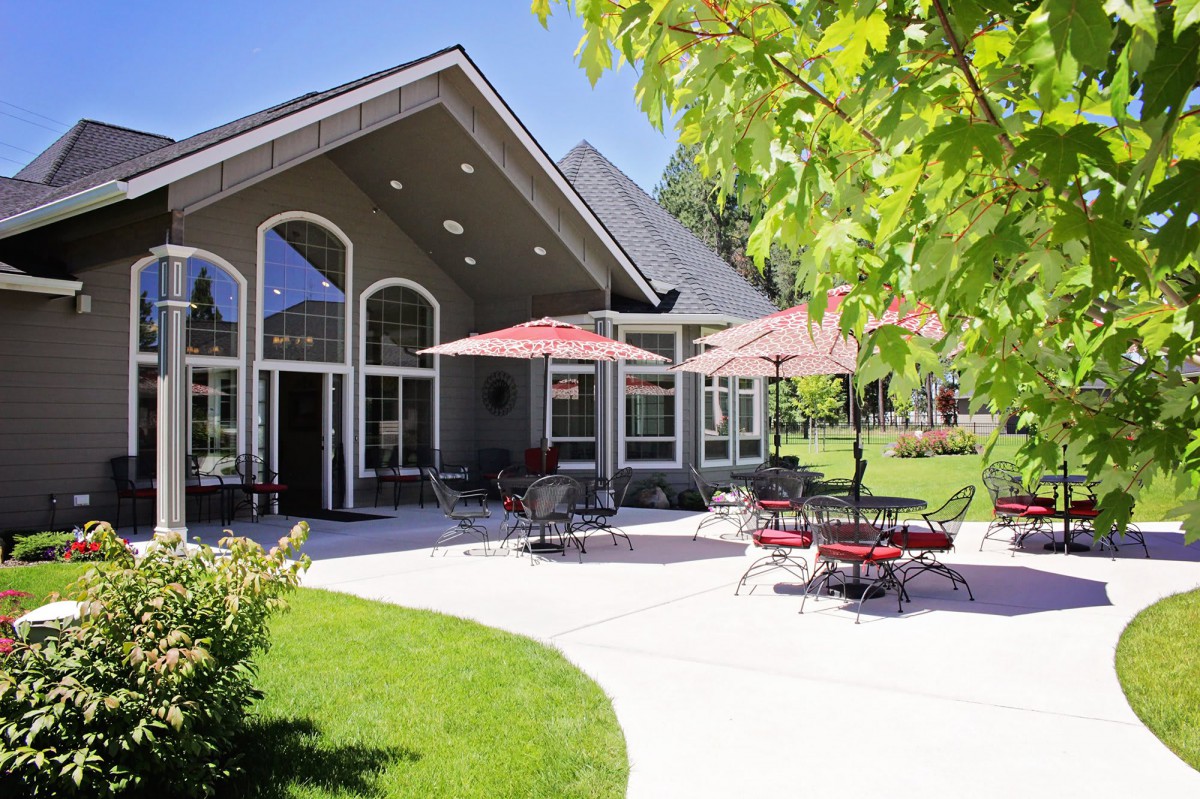 A modern house with a spacious patio featuring outdoor furniture and umbrellas, surrounded by a lush green lawn and trees in bright sunlight.