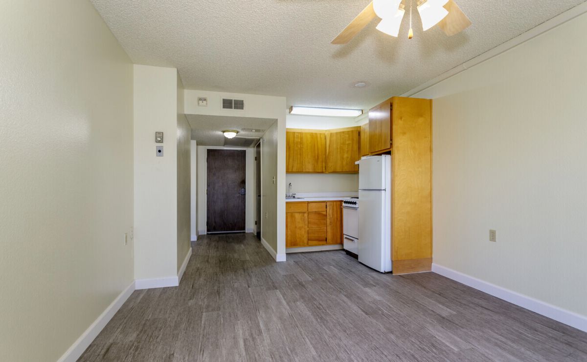 A view of an empty apartment with a small kitchen featuring wooden cabinets, a refrigerator, and hardwood flooring.