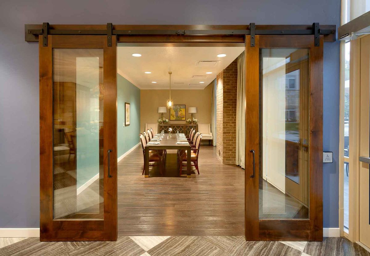 A modern dining room with wooden flooring, featuring a long table and chairs, illuminated by a chandelier and accessed through sliding glass doors.