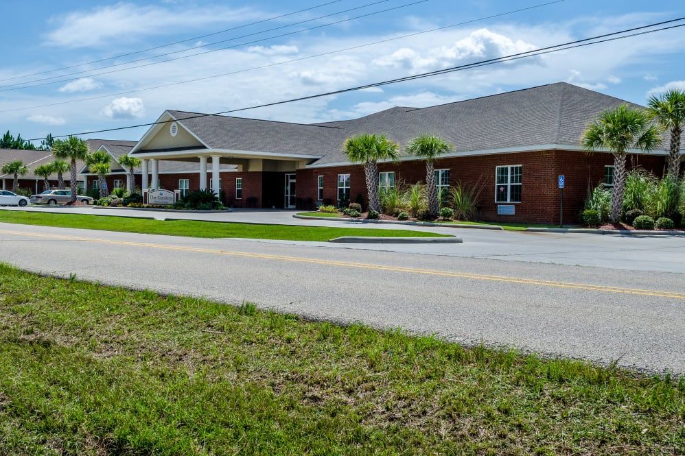 A one-story brick building with a covered entrance, situated alongside a road with palm trees and grassy surroundings under a partly cloudy sky.