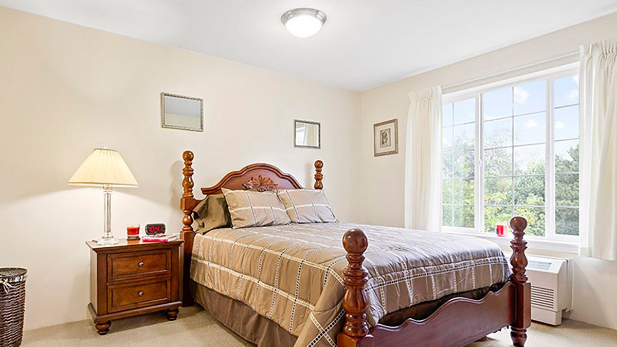 A cozy bedroom with a wooden bed and matching nightstand, complemented by neutral decor and plenty of natural light from a large window.