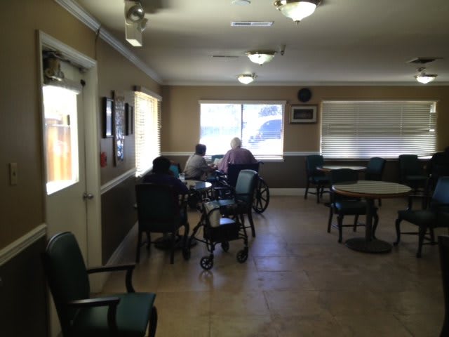 A calm nursing home common area with seniors sitting at tables near a window.