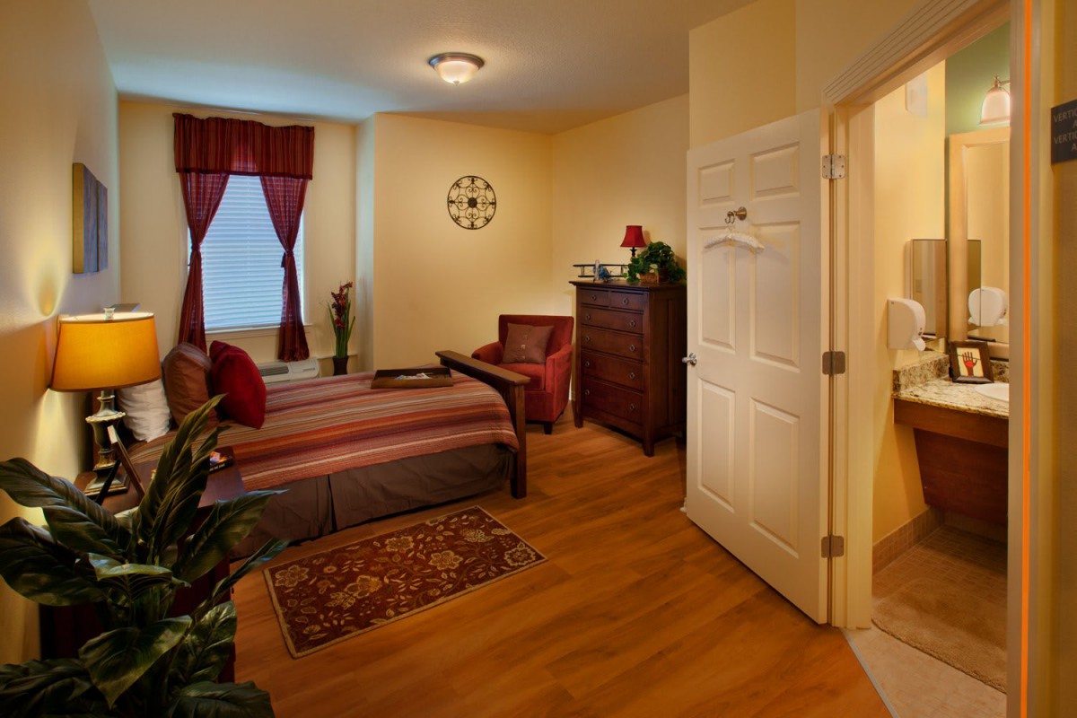 A cozy bedroom with warm lighting, featuring a bed, armchair, chest of drawers, and decorative elements.