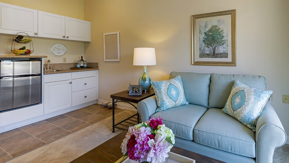 A cozy living room with a light blue sofa and decorative cushions, featuring a lamp on a side table and colorful flowers adding vibrancy to the space.