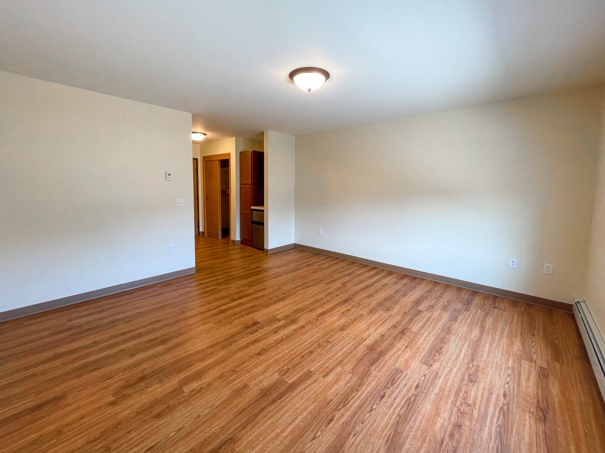 An empty room with hardwood flooring and a central light fixture, featuring beige walls and a doorway leading to another area.