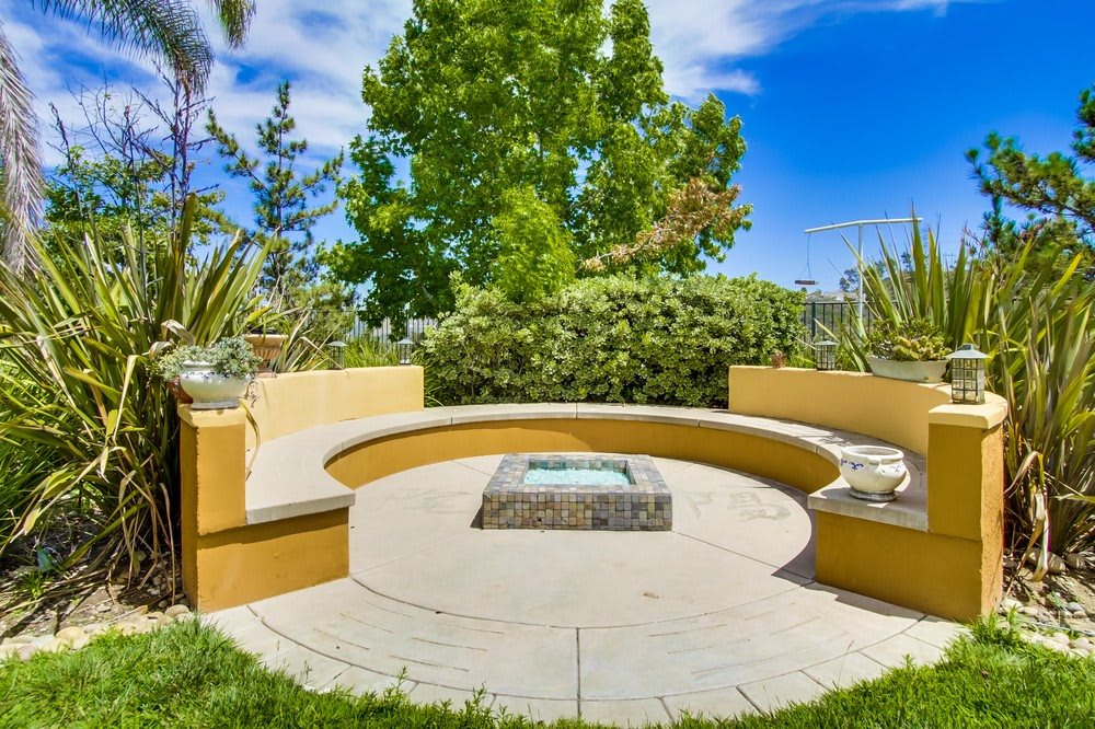 A sunny outdoor patio with a circular seating area and a central fire pit surrounded by lush greenery.