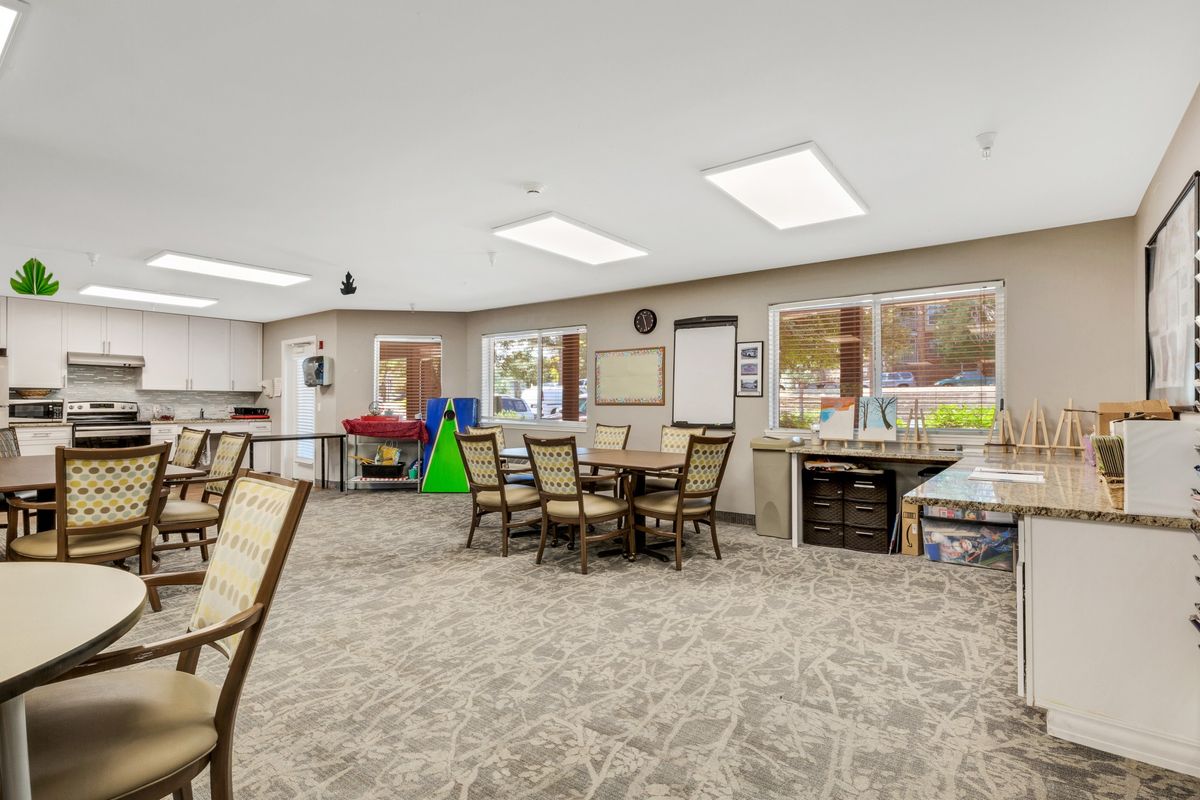 A spacious room featuring tables and chairs, with a carpeted floor, large windows, and overhead lighting.