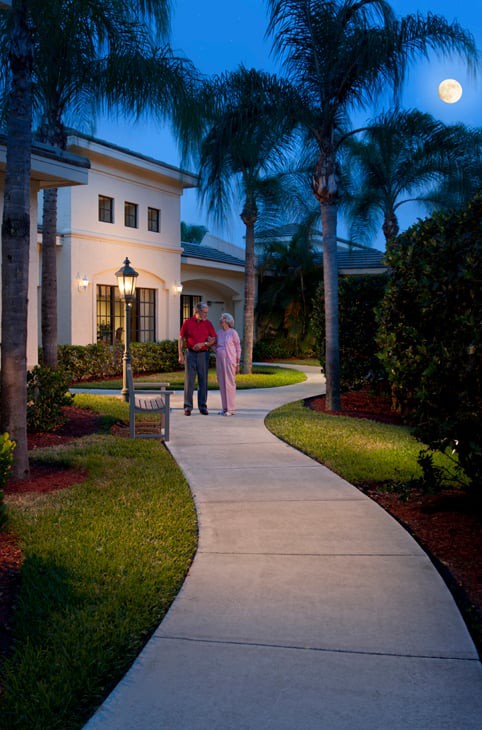 A couple stands on a beautifully lit pathway outside a modern house during the evening.