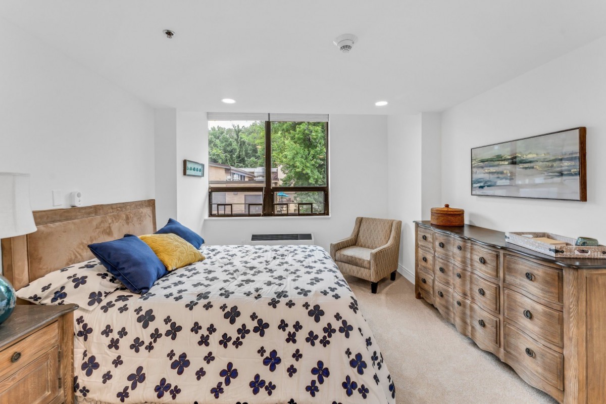 A cozy bedroom with a patterned bedspread, wooden furniture, and a large window allowing natural light.