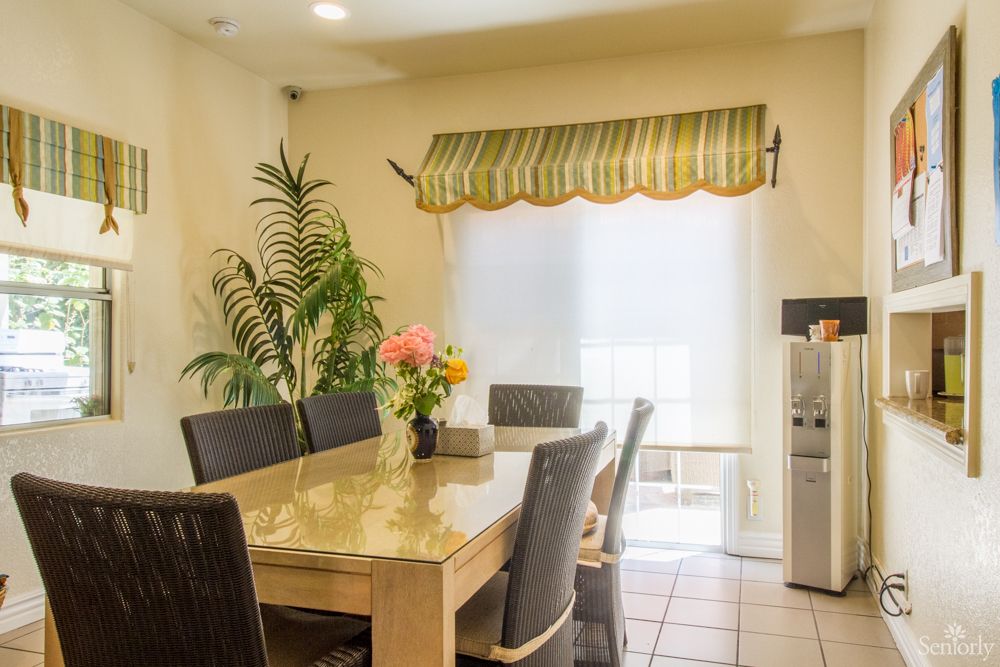 A cozy dining room with a table set, surrounded by chairs and decorated with a flower vase, plant, and window curtains.