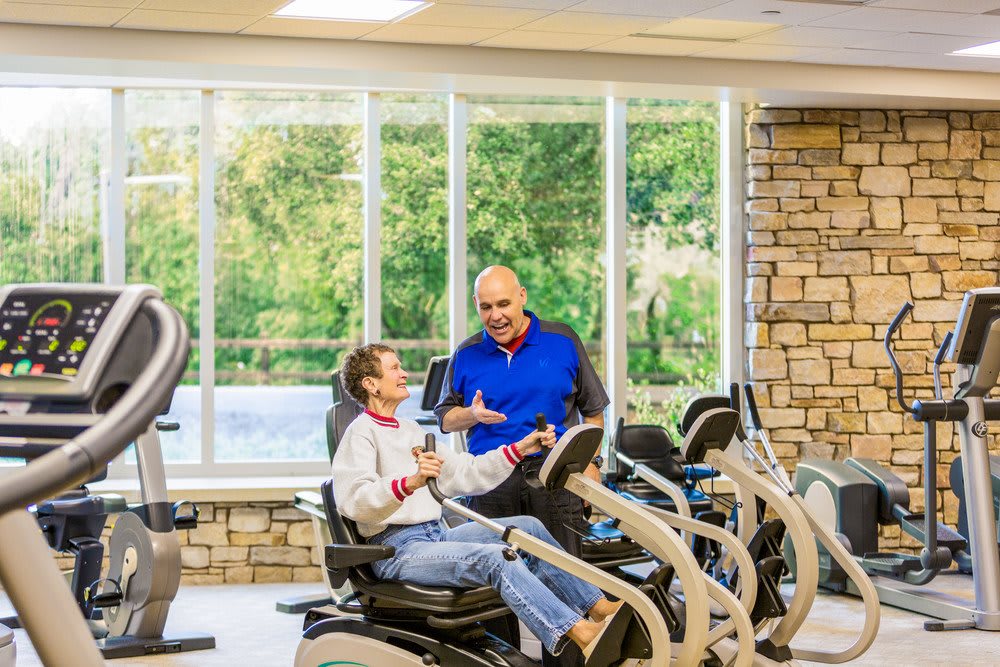 Two people engaging in conversation while using exercise equipment in a gym setting.