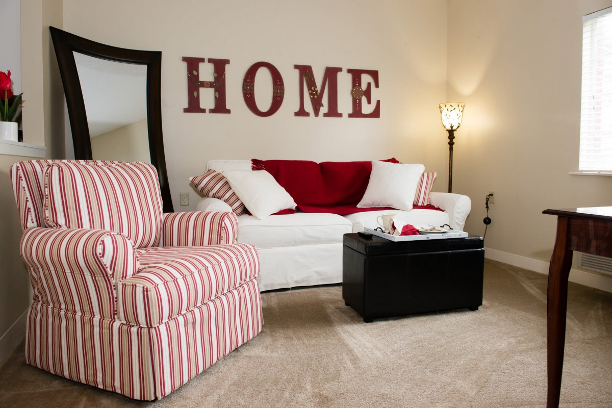 A cozy living room featuring a white sofa with red and white cushions, a striped armchair, modern lighting, and 'HOME' wall decor.