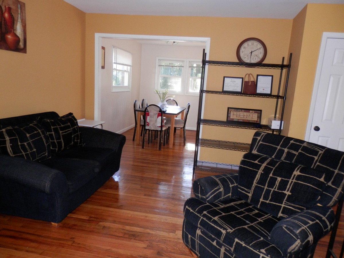 A cozy living room with a sofa set and an adjacent dining area, featuring wooden flooring and a shelving unit.