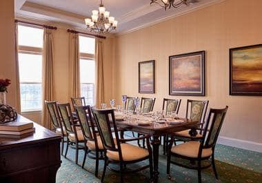 An elegant dining room with a large table set for a formal meal, surrounded by cushioned chairs and illuminated by chandeliers.