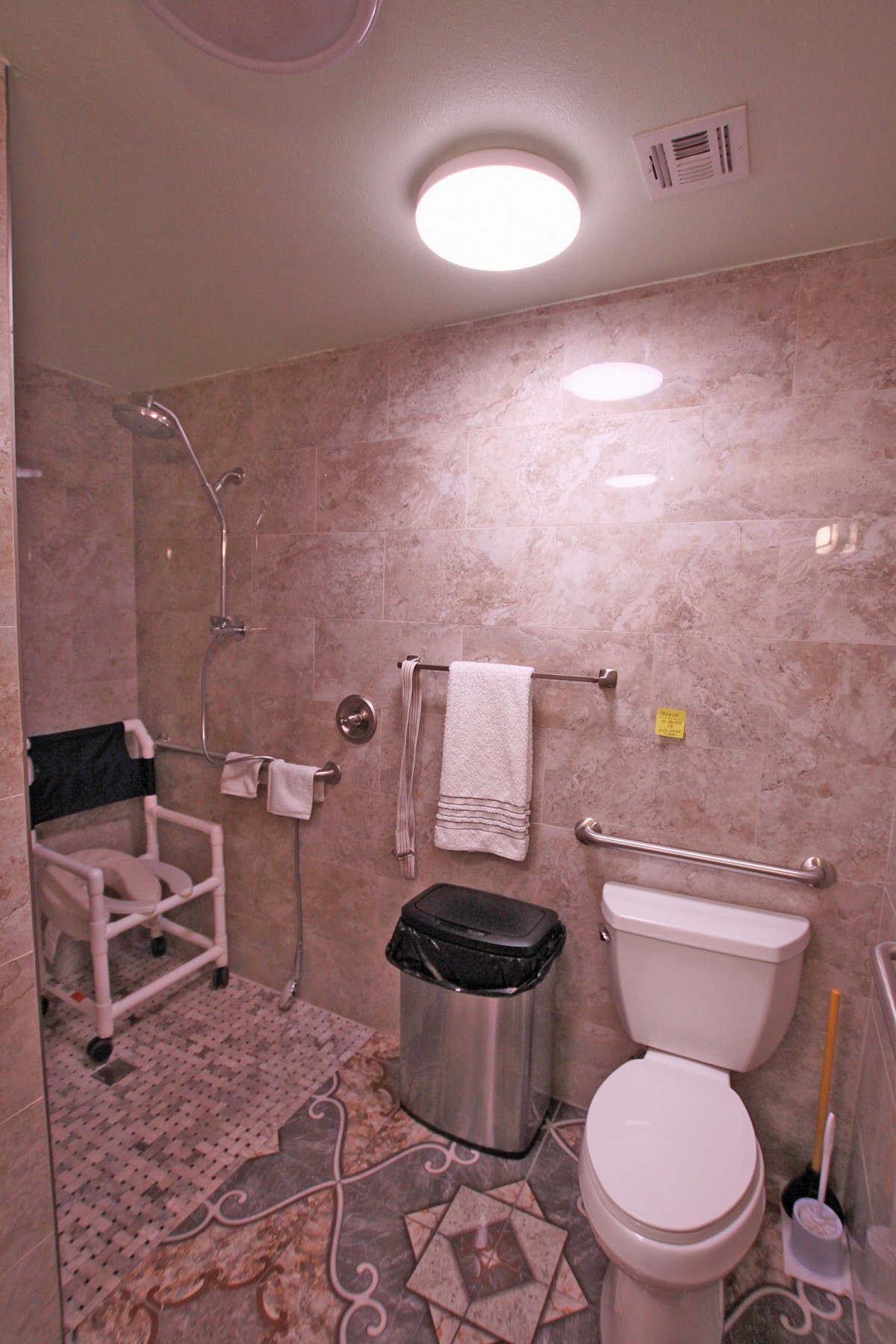 An accessible bathroom featuring a shower chair, toilet with handrails, and towel rack.