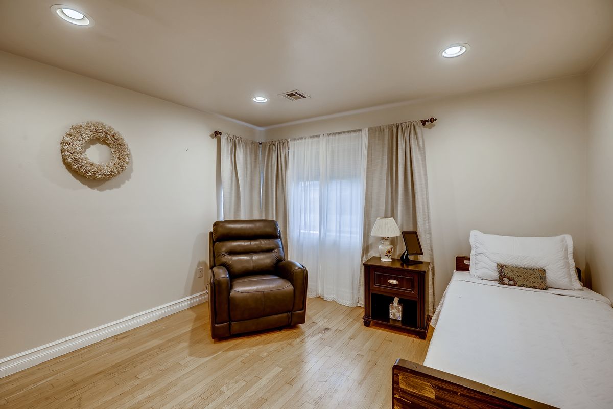 A cozy bedroom featuring a recliner chair, bed, nightstand with a lamp, and soft curtains.