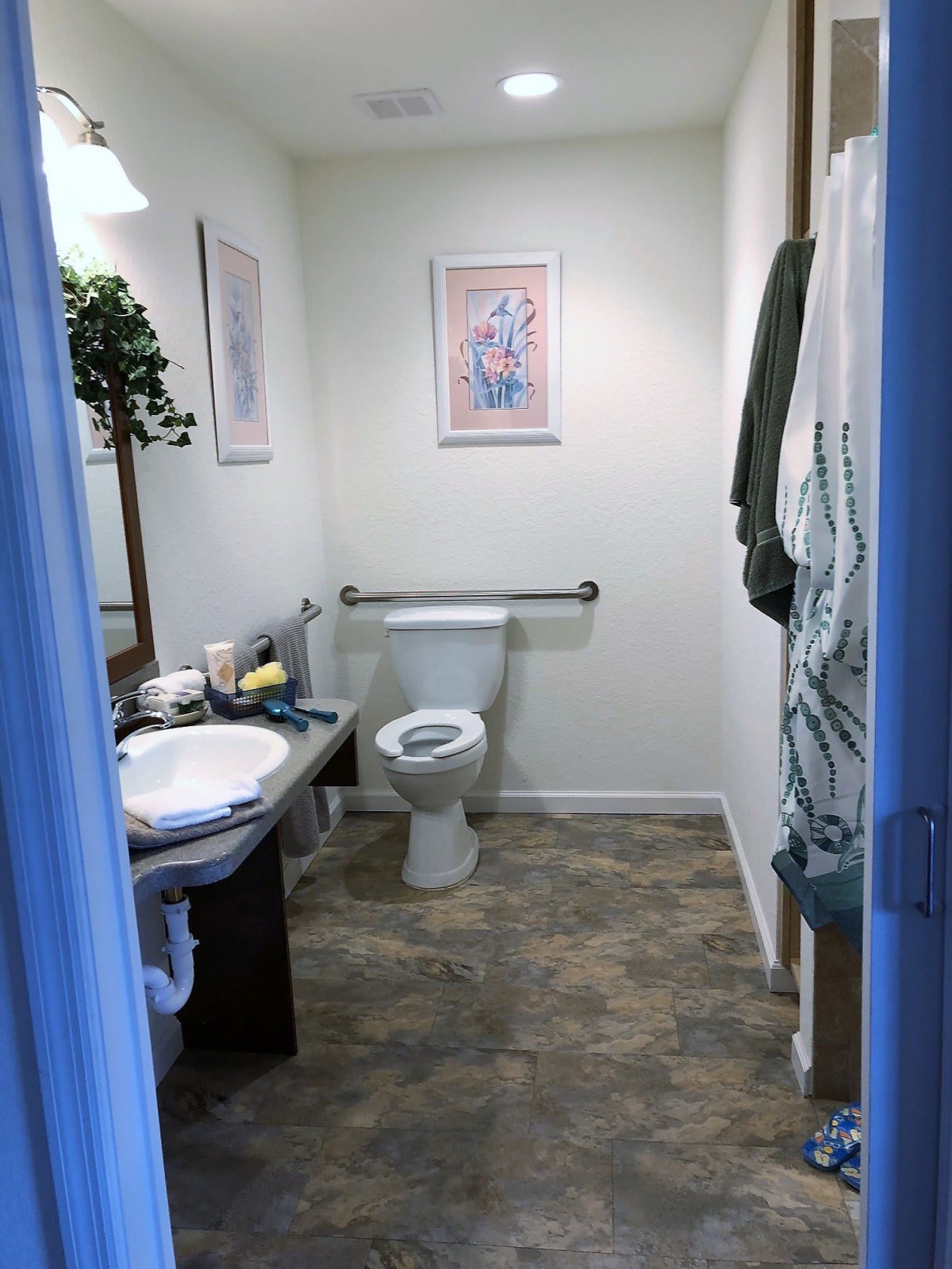 A small, neatly organized bathroom with a toilet, sink, mirror, and wall art.