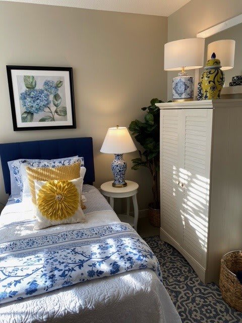 A cozy bedroom featuring a single bed with blue and white bedding, accented by yellow pillows. A lamp sits on a side table next to the bed, alongside decorative items and artwork on the walls. A tall dresser completes the room's furnishings.