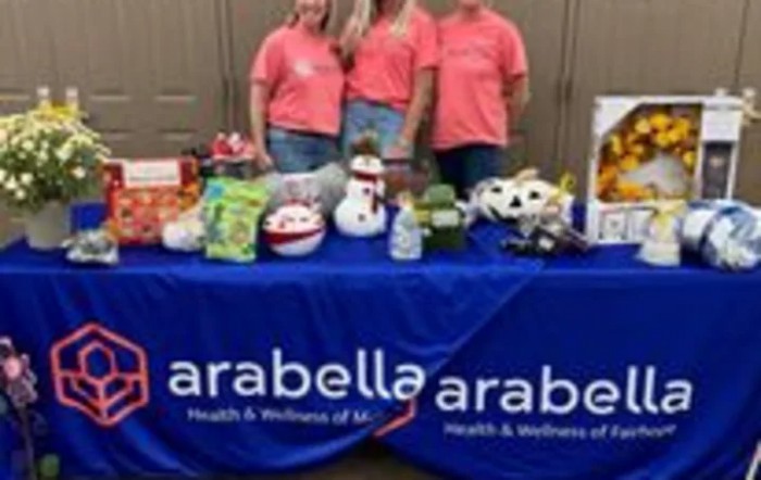 Three individuals standing behind a promotional table for Arabella Health & Wellness, showcasing various items.