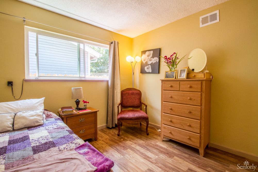 A cozy bedroom with wooden flooring, featuring a dresser and a chair near the window.