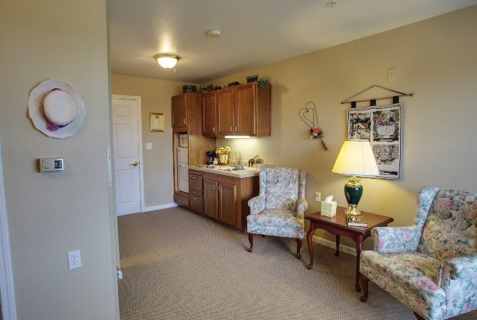 A cozy living area with a kitchenette, featuring two upholstered chairs and a small table with a lamp.