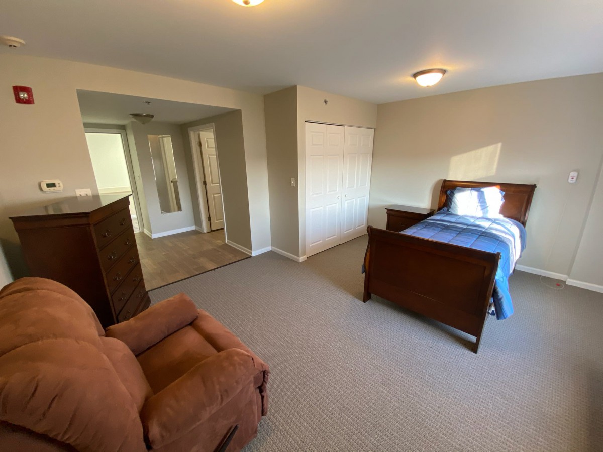 A spacious bedroom with a single bed, armchair, and dresser, featuring a carpeted floor and ample natural light.