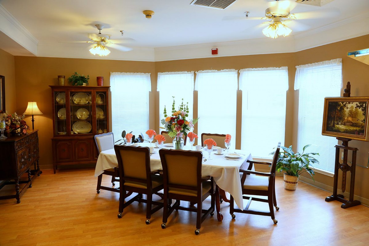 A well-lit dining room with a table set for six, featuring a floral centerpiece and surrounded by large windows.