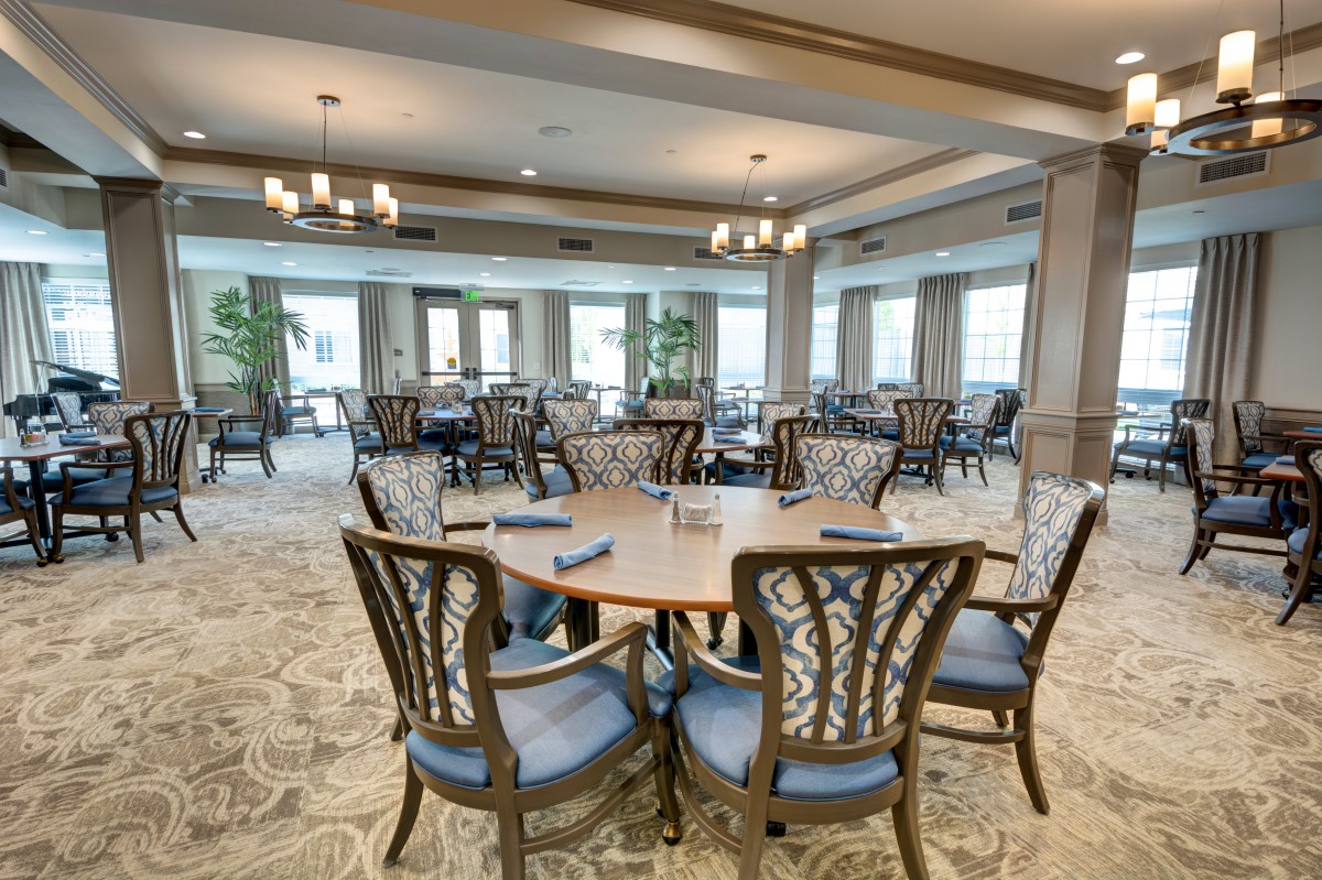 A spacious dining room with elegantly set tables and stylish chairs, featuring large windows and chandeliers.