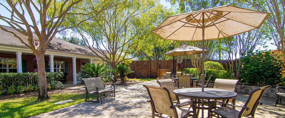 A sunny backyard patio with outdoor furniture and umbrellas surrounded by trees.