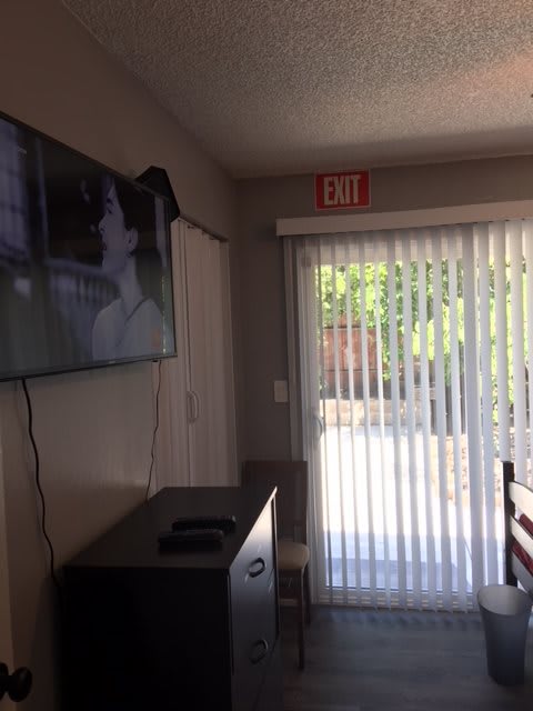 A living room with a wall-mounted television and a glass door leading outside, covered by vertical blinds.