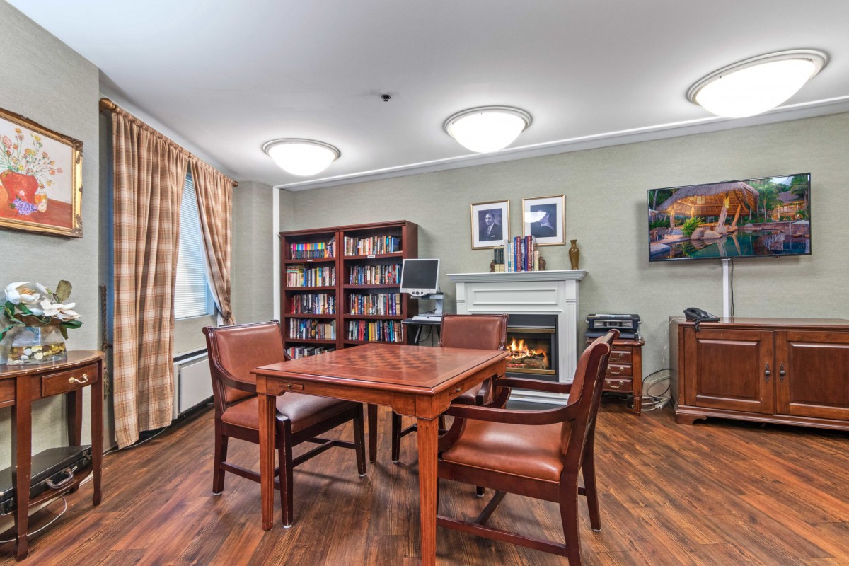 A cozy library room with a wooden table and chairs, surrounded by bookshelves and decorated with artwork above a fireplace.