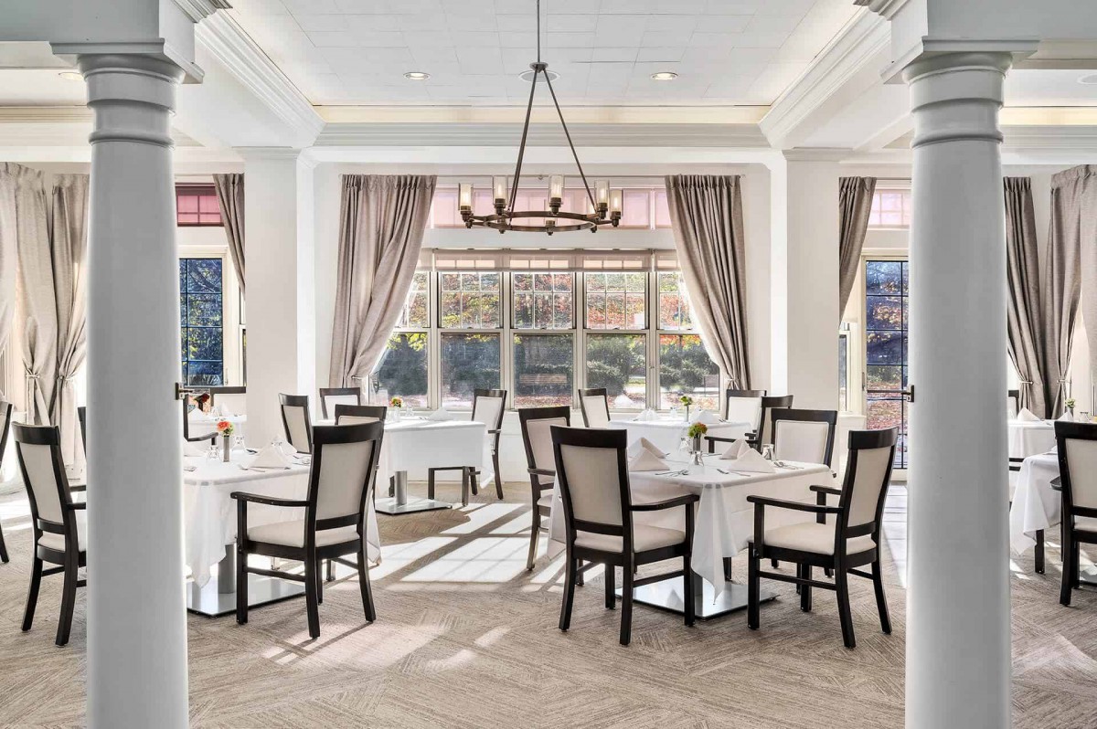 An elegant dining room with neatly arranged tables and chairs, complemented by a chandelier and large windows with curtains.