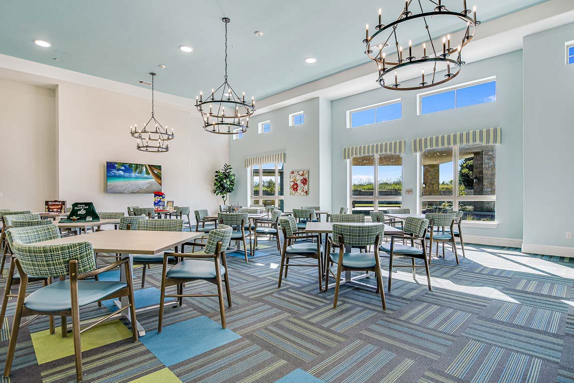 A modern dining area with multiple tables and chairs, illuminated by chandeliers and natural light from large windows.