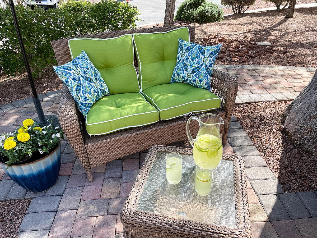 A cozy patio setup with green cushioned seating and a glass table holding a pitcher and glass of lemonade.