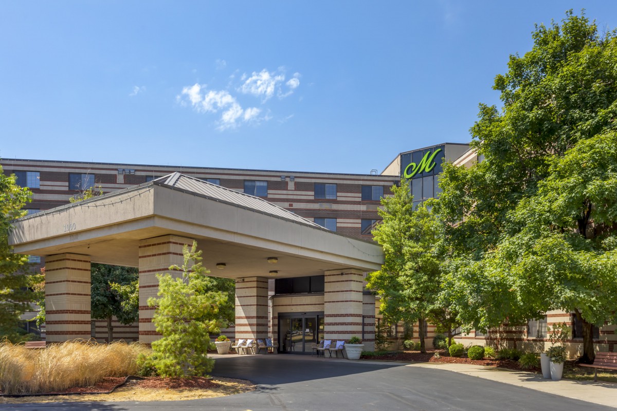 A modern hotel entrance with surrounding trees and a clear blue sky.