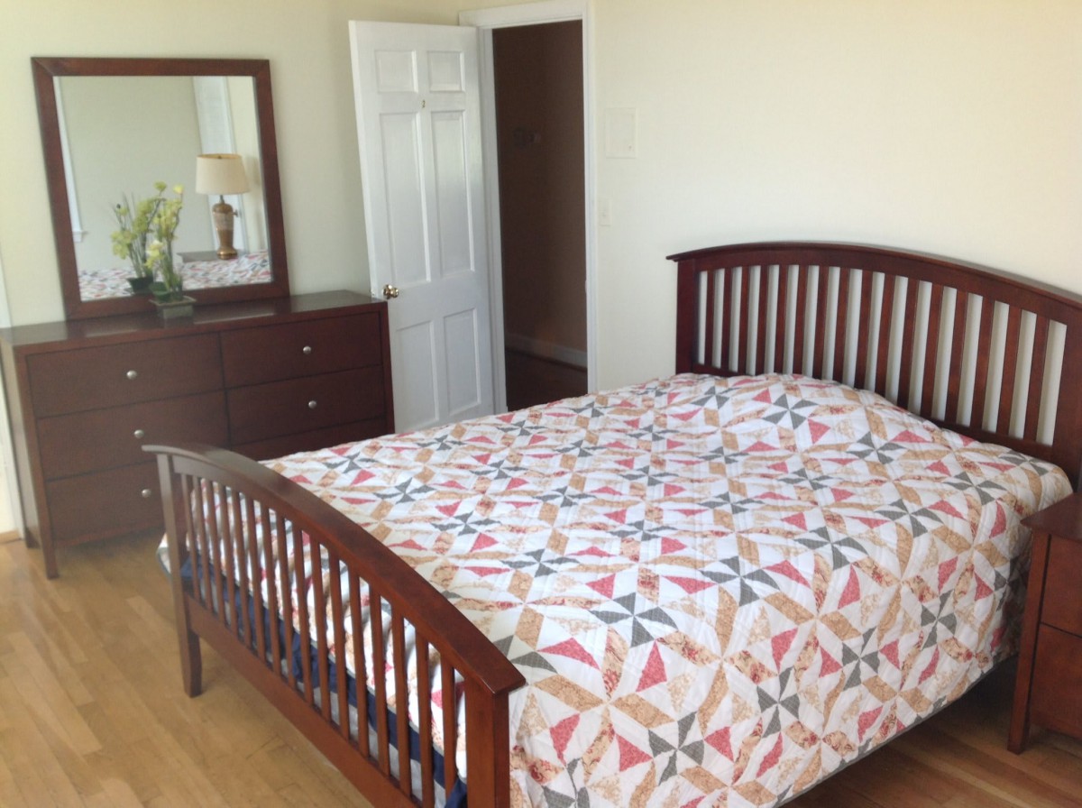 A tidy bedroom with a wooden bed frame and colorful quilt, complemented by a dresser with a mirror.