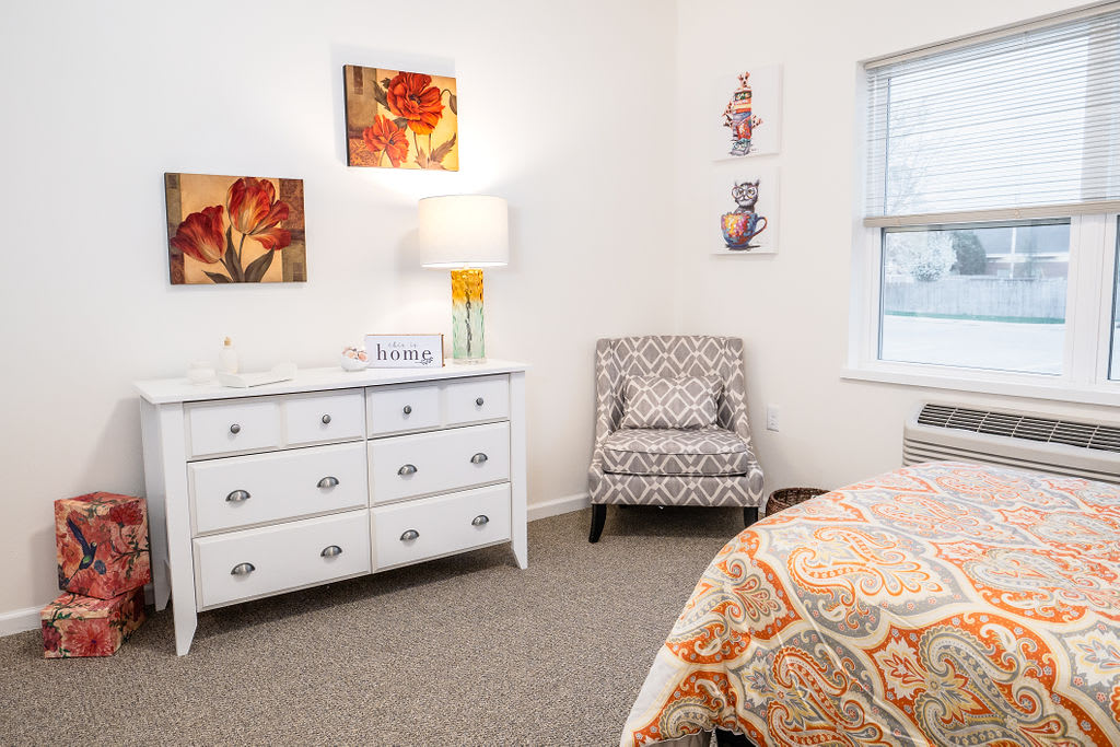 A cozy bedroom interior featuring a white dresser, patterned chair, and colorful artwork.