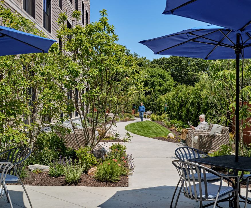A peaceful outdoor patio area with tables, chairs, and blue umbrellas surrounded by greenery and a garden pathway.