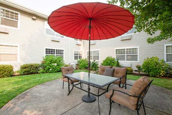 A patio with a glass table and chairs, shaded by a large red umbrella in a garden setting.