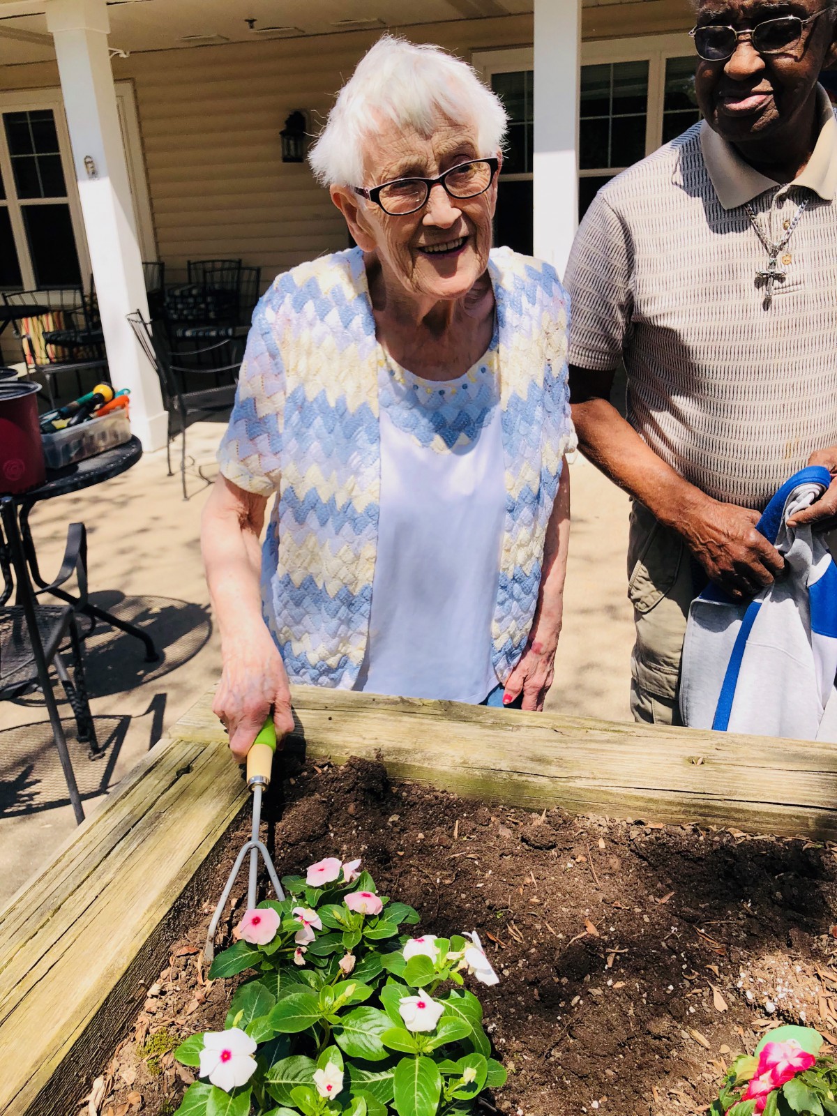 An elderly woman is happily gardening outdoors, planting flowers in a raised bed.