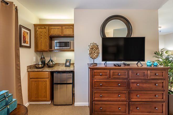 A cozy hotel room featuring a kitchenette with a microwave and mini fridge, a wooden dresser topped with a television, and a decorative mirror above.