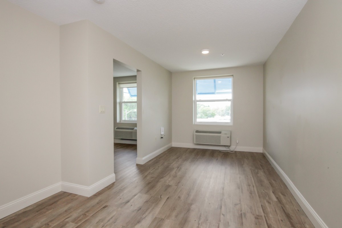 An empty, well-lit living room with wooden flooring and a window.