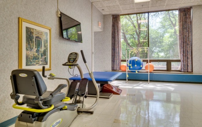 A fitness room with exercise equipment, including a stationary bike and therapy table, next to a large window.