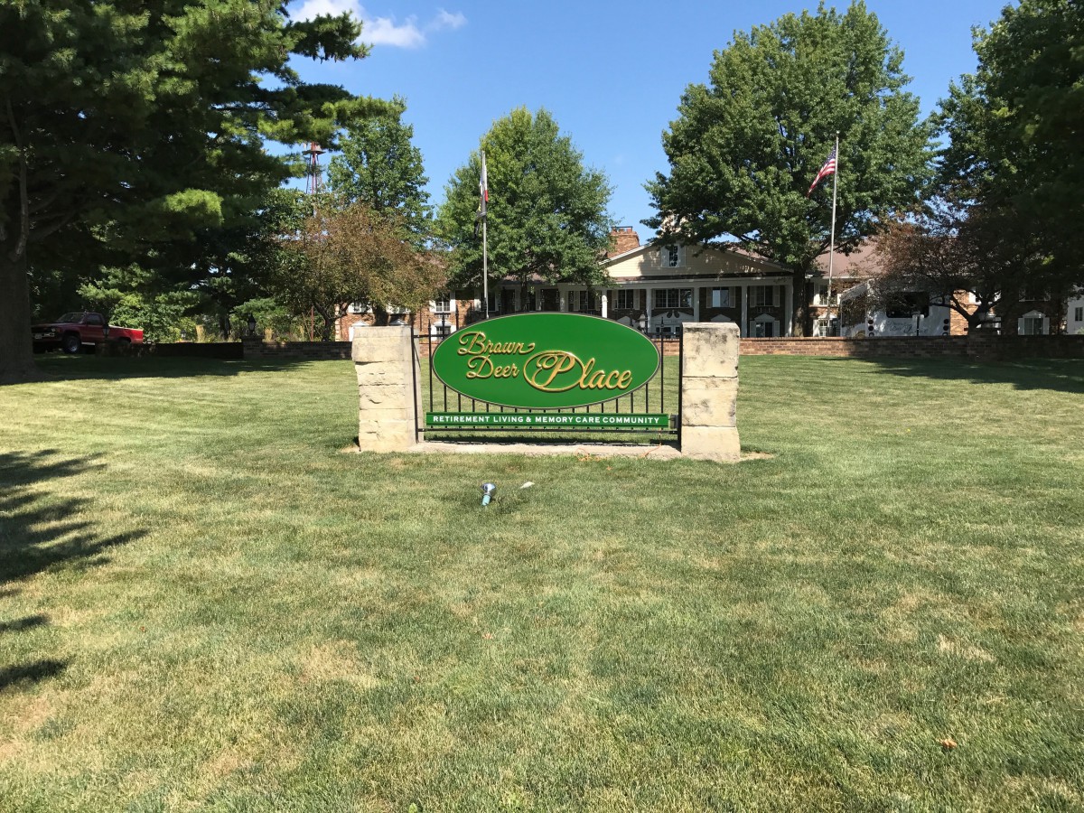 A welcoming green sign on a neatly maintained lawn with trees and a building in the background.