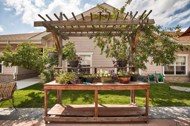 A backyard garden with a wooden pergola and a potting table surrounded by lush greenery on a sunny day.