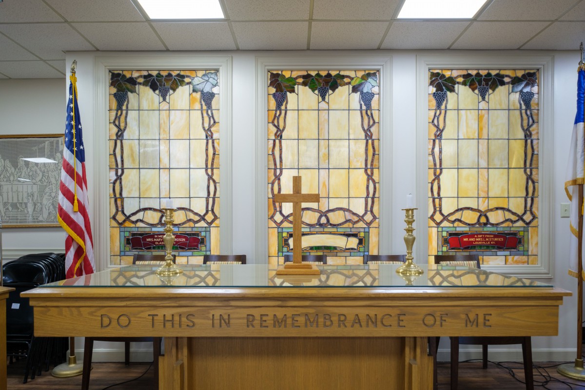 A church altar with a communion table adorned with a cross and candles, set against stained glass windows.