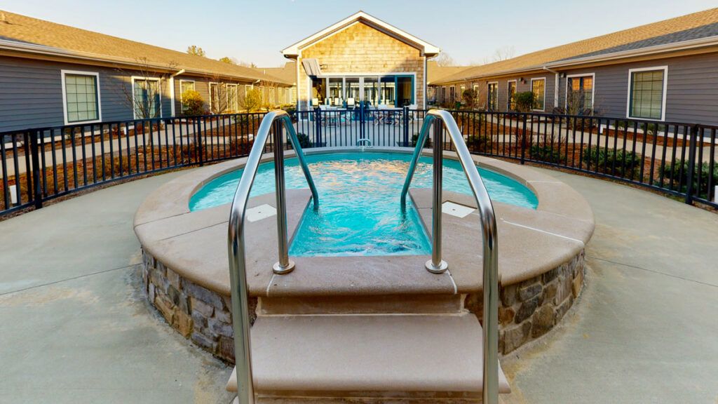A small circular outdoor pool with metal steps and railing, surrounded by a fence, situated near a building during the day.