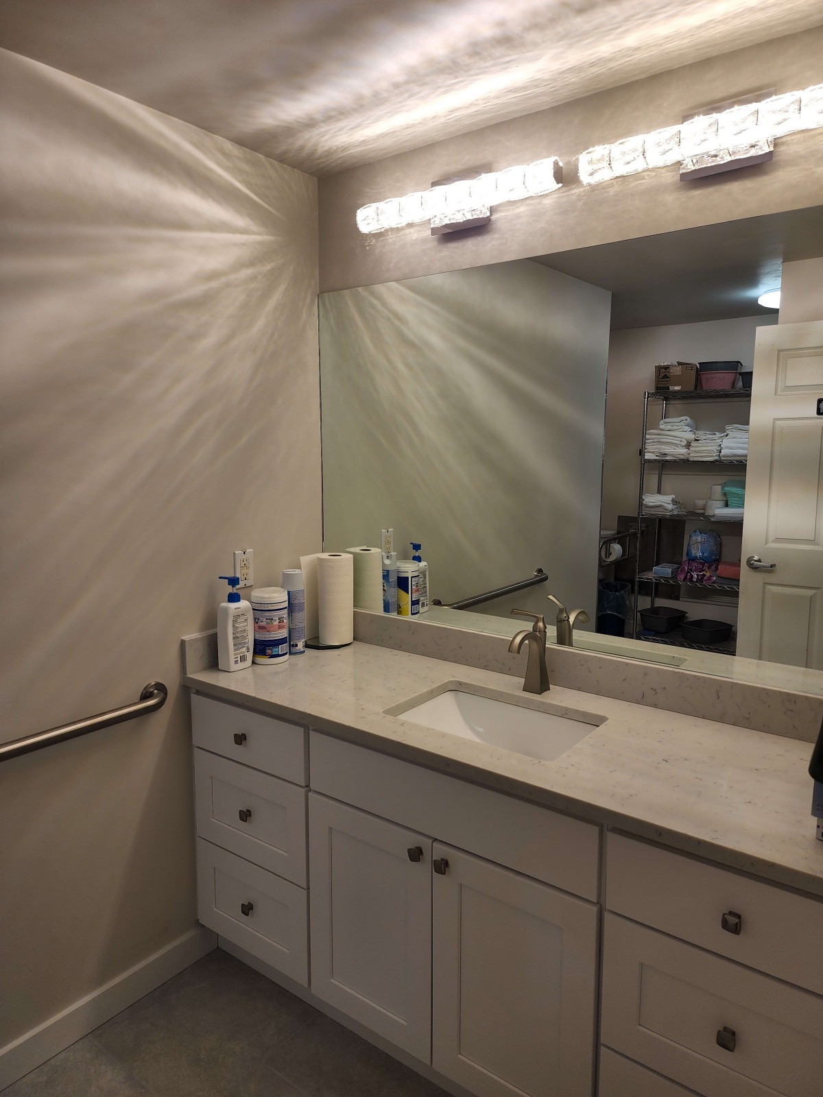 A clean bathroom with a large mirror, countertop sink, and cabinets beneath. It has overhead lighting and various hygiene products on the counter.