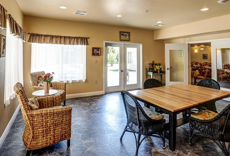 A cozy living and dining area featuring wicker furniture, a wooden table, and ample natural light from large windows.