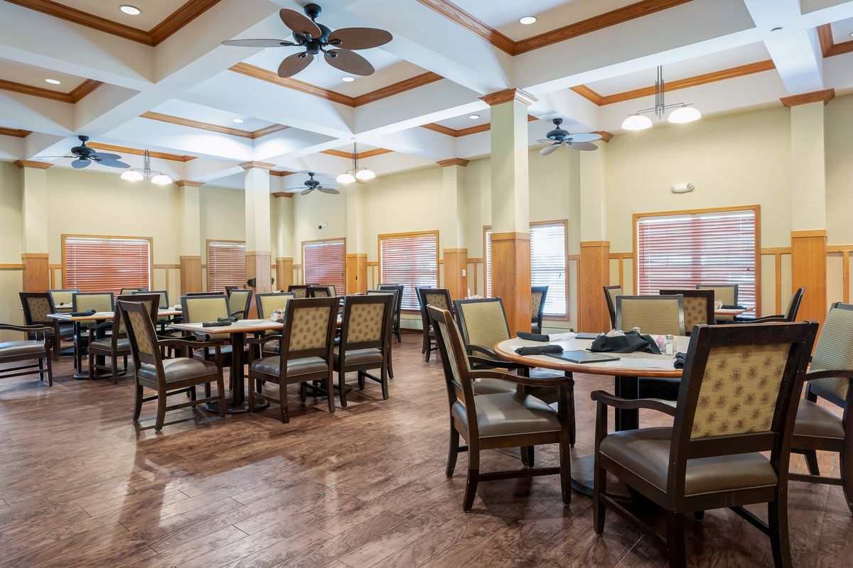 An empty dining hall with neatly arranged tables and chairs, featuring ceiling fans and a wooden floor.